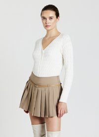 Weiße, strukturierte Strickjacke mit tiefem V-Ausschnitt, kombiniert mit einem beigefarbenen, gefalteten Minirock, der mit einem beigen Gürtel getragen wird, sowie kniehohen weißen Stiefeln.