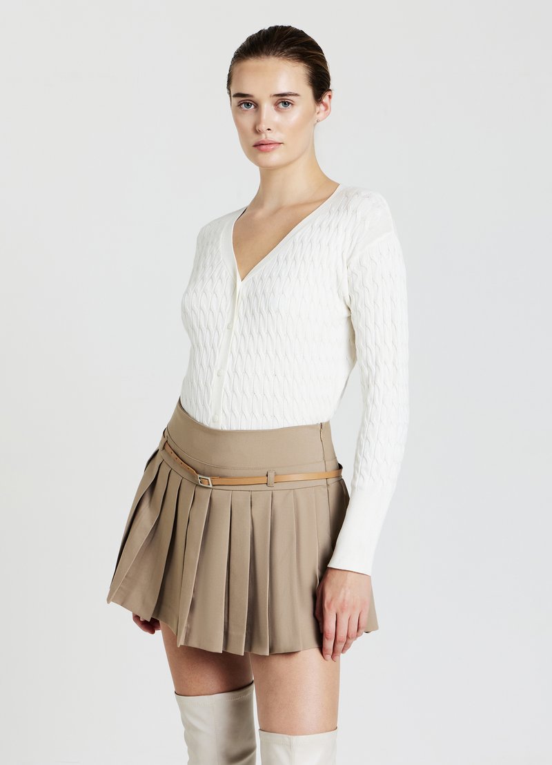 Weiße, strukturierte Strickjacke mit tiefem V-Ausschnitt, kombiniert mit einem beigefarbenen, gefalteten Minirock, der mit einem beigen Gürtel getragen wird, sowie kniehohen weißen Stiefeln.