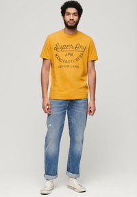 T-shirt di cotone giallo senape con grafica "Super Dry", abbinata a jeans denim chiari strappati, con risvolti, e sneakers bianche.