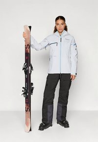 Femme en veste de ski gris clair, pantalon de ski noir et bottes, debout en tenant une paire de skis verticaux avec fixations sur un fond blanc.