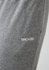 Graue Stoff-Jogger mit glatter Textur, Taschen an den Seiten und "SHOCKS" in Weiß auf dem linken Oberschenkel gestickt.