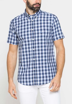 Uomo che indossa una camicia a maniche corte con motivo a quadri blu e bianco e pantaloni bianchi, in piedi davanti a uno sfondo semplice.
