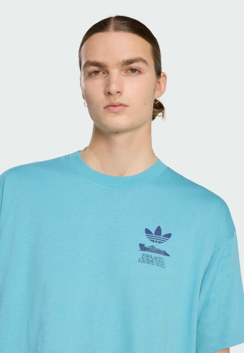 Giovane con i capelli lunghi raccolti che indossa una T-shirt Adidas azzurra chiara con logo e grafica sul petto.
