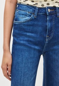 Sötétkék farmernadrág középmagas derékkal, narancssárga varrással, elülső zsebekkel és cipzáras sliccel. Simább, texturált szövet.