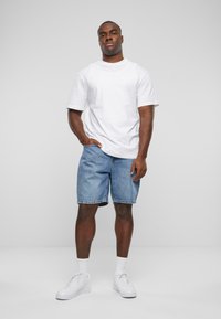 T-shirt blanc à manches courtes, shorts en denim bleu, baskets blanches et chaussettes. La tenue présente une coupe décontractée et une texture en denim léger.