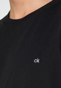 Czarna koszulka z okrągłym dekoltem z małym białym logo "ck" wyszytym na lewej piersi, noszona przez osobę.