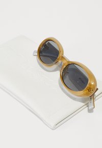 Des lunettes de soleil de couleur ambre avec des verres ronds surdimensionnés, présentées dans un étui blanc doux embossé avec "Le Specs". Les verres sont foncés avec un léger dégradé.