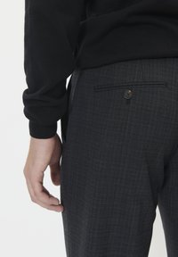 Sötét kockás nadrágot viselő férfi, hátul gombos zsebbel, fekete hosszúujjú pólóban, karja lazán a teste mellett egy egyszínű háttér előtt.