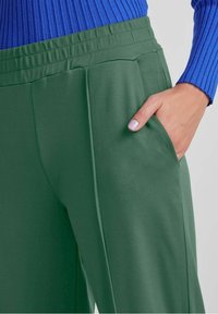 Zielone, wysokie spodnie o gładkiej fakturze, z bocznymi kieszeniami i luźnym krojem. Połączone z niebieską, prążkowaną bluzką.