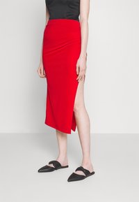 Roter Midikleid mit einem seitlichen Schlitz, kombiniert mit schwarzen flachen Mules. Glatte Textur und figurschmeichelndes Design, das die Silhouette betont.