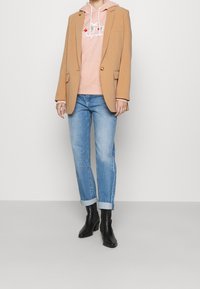 Beżowa marynarka na różowej bluzie z kapturem z logo, niebieskie podwinięte dżinsy i czarne botki na obcasie. Gładkie faktury i dopasowany krój.