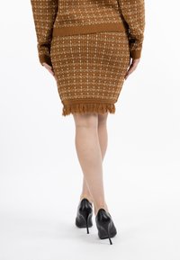 Fustă tricotată maro cu un model texturat, detalii cu tiv dezhicat, purtată cu pantofi cu toc înalt negri. Fundalul este alb simplu.