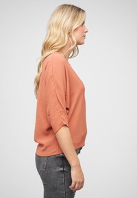 Licht koraalkleurige oversized blouse met een gestructureerde afwerking en een losse pasvorm. Heeft mouwen tot op de elleboog en een ronde halslijn. Gekenmerkt door een combinatie met donkergrijze jeans.