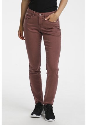 Modèle portant un pantalon marron ajusté avec des poches avant, associé à des baskets noires et un haut noir, debout devant un fond blanc.