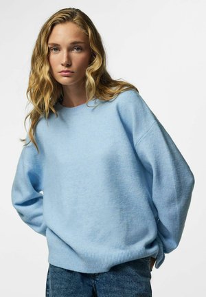 Jeune femme aux longs cheveux blonds ondulés, portant un pull oversize bleu clair et un jean bleu, debout devant un fond blanc uni.