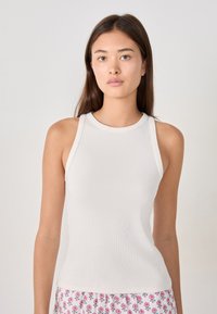 Young woman with long brown hair wearing a sleeveless white ribbed top and floral patterned pants against a plain light background.