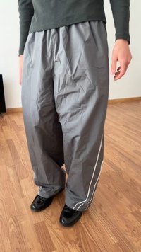 Pelēki, krunkaini, platā kāju bikses ar elastīgu jostu un baltām sānu svītrām, apvienotas ar melniem apaviem uz koka grīdas.