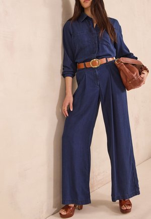 Femme portant une combinaison bleu foncé à jambes larges avec manches retroussées, ceinture marron, sandales compensées marron et tenant un sac à main en cuir marron.