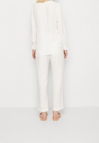 Chemise blanche à manches longues avec un ourlet arrondi, associée à un pantalon blanc ample. Des sandales à design tissé complètent la tenue.