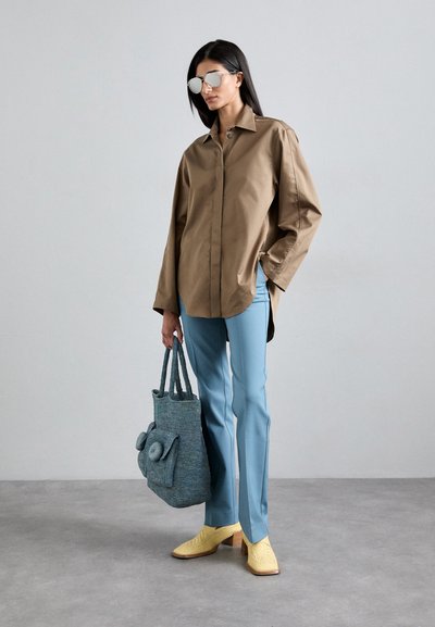 Brown oversized shirt, light blue tailored pants, textured blue tote bag, yellow heeled shoes, and reflective sunglasses.