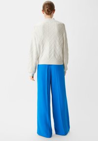 Weißer gestrickter Pullover mit einem strukturierten Zickzackmuster, kombiniert mit leuchtend blauen, weit geschnittenen Hosen. Der Pullover hat gerippte Bündchen und Saum.