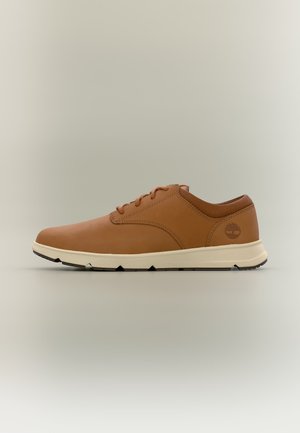 Chaussure décontractée en cuir marron à lacets avec semelle blanche et logo embossé sur le talon, vue de côté sur fond neutre.