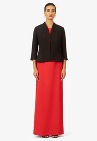 Zwarte gestructureerde jas met driekwart mouwen over een lange rode jurk. Glad stof met een getailleerd silhouet en minimale details.