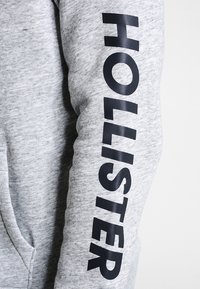Felpa grigia con una texture morbida, con la scritta "HOLLISTER" stampata in lettere nere in grassetto lungo la manica. Design classico, vestibilità casual.