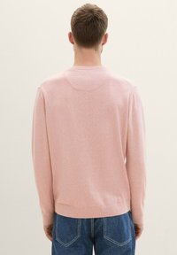 Svetlo roza pleteni pulover z rebrastimi manšetami in obrobo, z zaobljenim izrezom na hrbtni strani. Nosi se z modrimi džinsami.