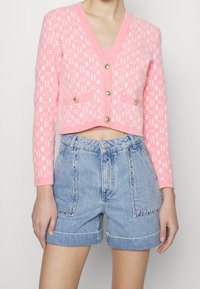 Różowy krótki kardigan z teksturowanym geometrycznym wzorem i złotymi guzikami, w połączeniu z jasnoniebieskimi szortami dżinsowymi z nitowanymi akcentami.