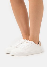 Hvite lær low-top joggesko med rund tå, teksturert overflate, flate snører og kunstlærdetalj ved hælen, satt mot en ensfarget bakgrunn.
