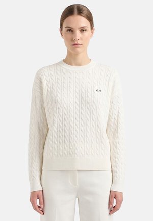 Giovane donna con capelli lisci indossa un maglione bianco a trecce e pantaloni bianchi, in piedi contro uno sfondo chiaro e semplice.