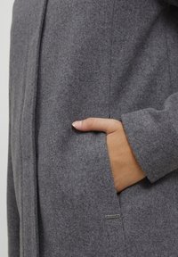Szary wełniany płaszcz o gładkiej fakturze, z boczną kieszenią i małą metką z logo. Projekt zawiera czyste linie i kobiecą sylwetkę.