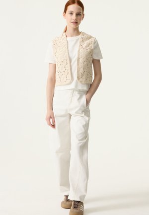 Chaleco beige de crochet sobre una camiseta blanca, combinado con pantalones de pierna ancha de color claro y zapatillas marrones. Diseño sencillo con motivos florales.