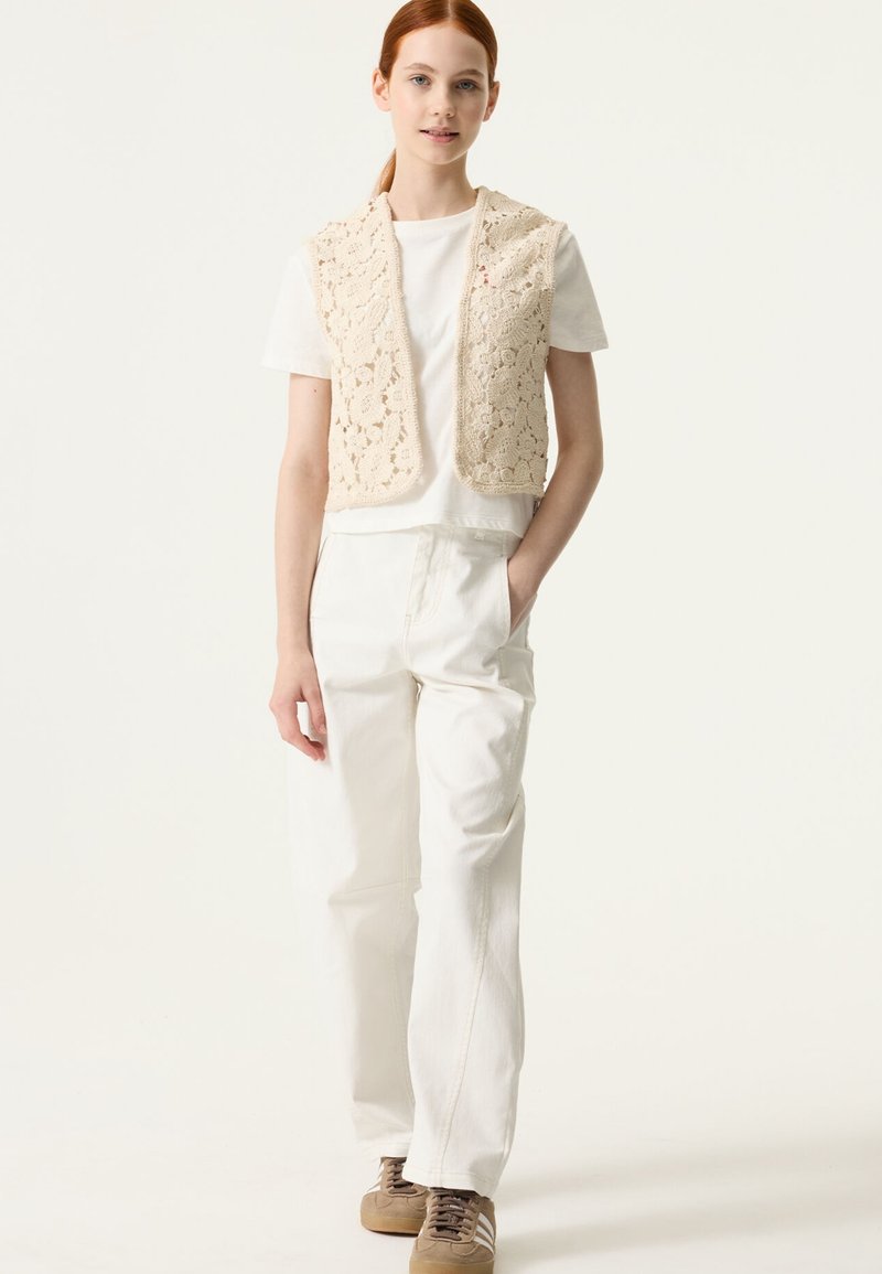 Veste beige en crochet portée sur un t-shirt blanc, associée à un pantalon à jambes larges de couleur claire et des baskets marron. Design simple avec des motifs floraux.