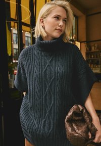 Wolliger, dunkelblauer Rollkragenpullover mit Zopfmuster und kurzen Ärmeln, kombiniert mit einer strukturierten, braunen, gewebten Handtasche.