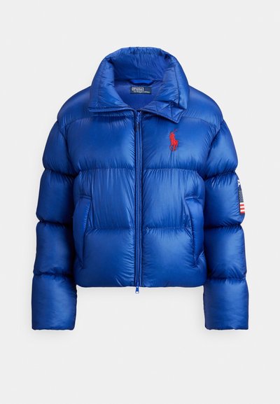 Blue puffer jacket featuring a high collar, quilted design, zip closure, and red logo. Includes American flag patch on the sleeve.