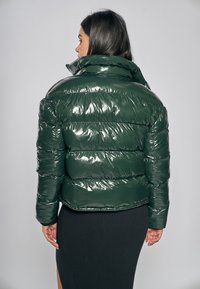 Blank mörkgrön puffjacka med hög krage och horisontellt quiltat mönster. Bakifrån ses en kortare stil och slät textur.