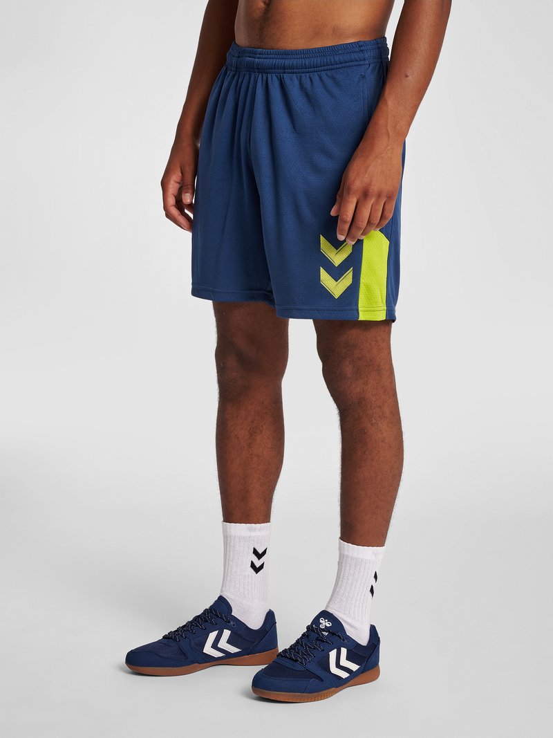 Mężczyzna noszący granatowe krótkie spodenki sportowe z neonowożółtym wzorem w jodełkę, białe skarpetki z czarnymi jodełkami i pasujące granatowe trampki.