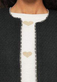 Kraveområdet viser en sort tekstureret jakke med perlekant over en hvid sweater med to guldglitrende hjerte-designs.