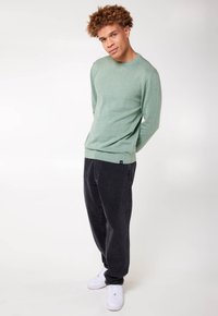 Maglione lungo verde chiaro a maniche lunghe con scollo rotondo, abbinato a pantaloni neri larghi e sneakers bianche, con una finitura leggermente texture.