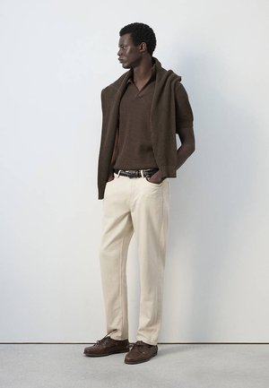 Un homme se tient debout en regardant à gauche, portant un pull marron avec un tricot assorti drapé sur les épaules, un pantalon crème et des chaussures marron contre un mur blanc.