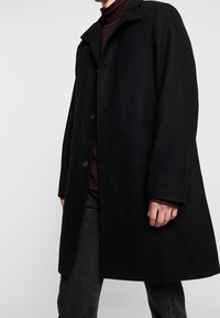 Musta villasekoitekot, jossa on suora leikkaus, napitus ja korkea kaulus. Yllä on maroon-värinen raidallinen poolopaita ja tummat farkut.