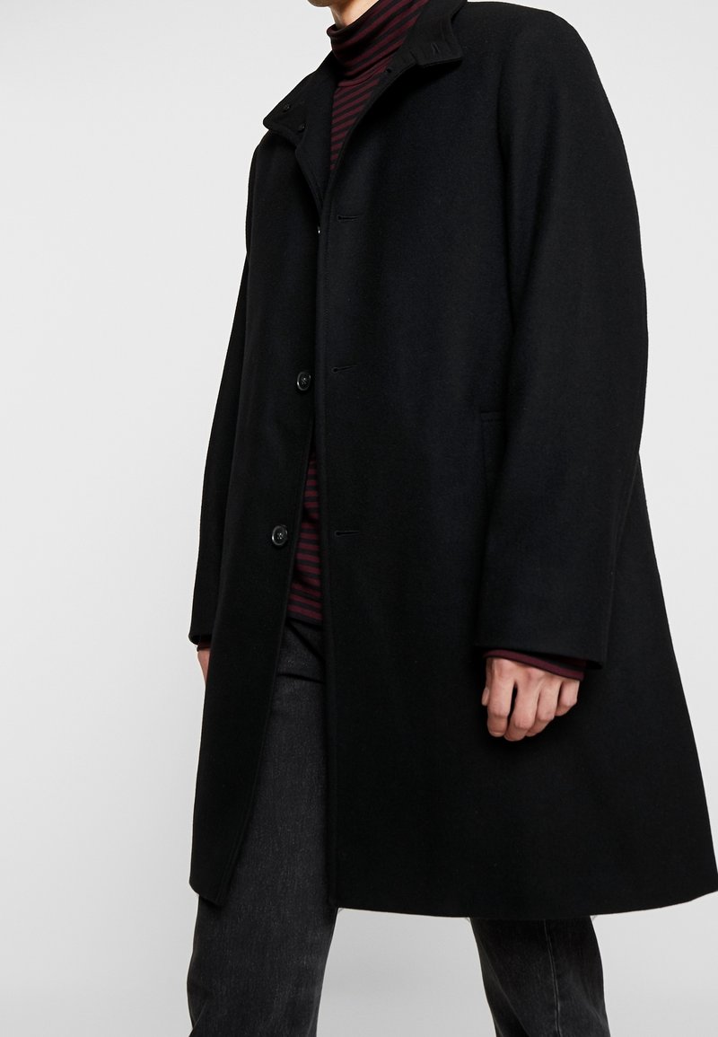 Musta villasekoitekot, jossa on suora leikkaus, napitus ja korkea kaulus. Yllä on maroon-värinen raidallinen poolopaita ja tummat farkut.