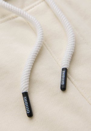 Zbliżenie na kremową tkaninę bluzy z kapturem, pokazujące biały pleciony sznurek z czarnymi plastikowymi końcówkami oznaczonymi "OMBRE."