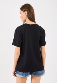 Tricou negru din bumbac cu o croială lejeră, cu mâneci scurte și guler rotund, asociat cu pantaloni scurți din denim. Prezentat din spate.