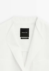 Bílý blazer s klopami, texturovanou tkaninou, přední kapsou a černou značkou s nápisem "Massimo Dutti" a "Vyrobeno v Portugalsku."