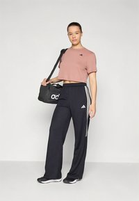 Rózsaszín, rövidített rövid ujjú póló és fekete nadrág fehér csíkokkal; a modell fekete duffle táskát hord; az Adidas logó látható a ruházaton és a táskán.