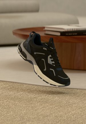 Črno-sivi atletski copat, ki visi nad bež preprogo iz tkanine, v ozadju pa lesen miza in knjige.