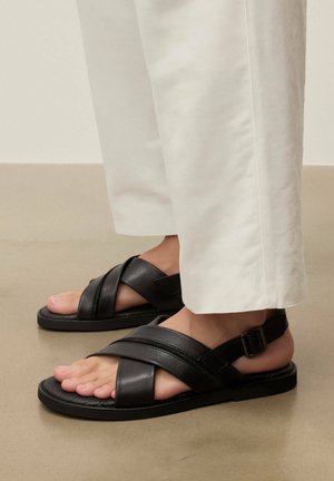 Fødder iført sorte lædersandaler med krydsede remme og spænder, kombineret med hvide bukser, stående på en glat gulv.